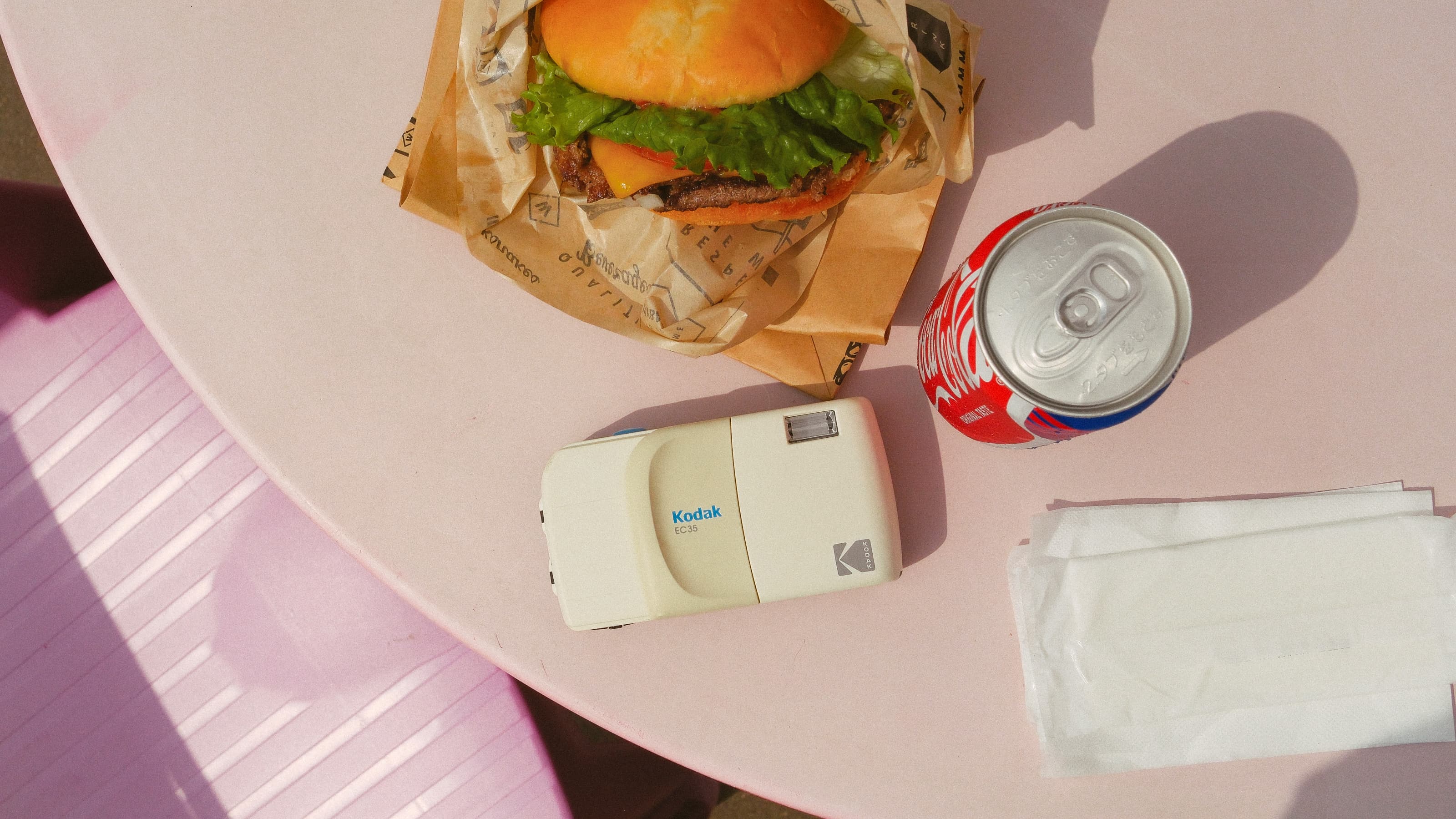 Foto de la cámara de película Kodak EC35 en blanco sobre una mesa junto a una hamburguesa y una lata de Coke