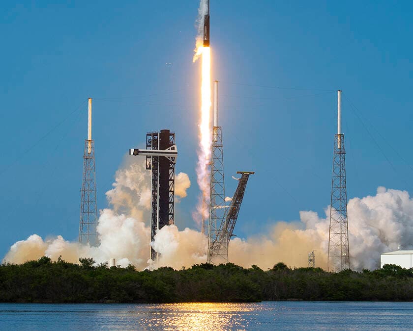 Falcon 9 rocket launches over the river, with the full rocket body and launch platform visible